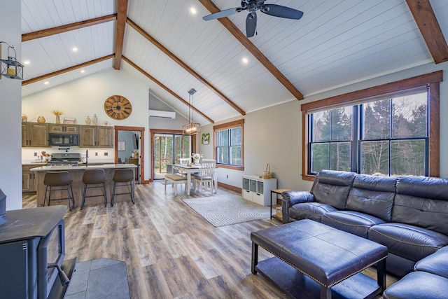living area with high vaulted ceiling, beamed ceiling, a wood stove, a ceiling fan, and light wood finished floors