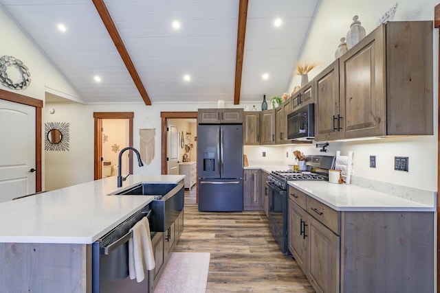 kitchen with vaulted ceiling with beams, light wood-style flooring, a center island with sink, appliances with stainless steel finishes, and a sink