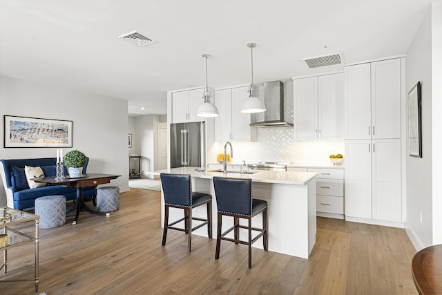kitchen featuring light countertops, an island with sink, high end refrigerator, wall chimney exhaust hood, and visible vents