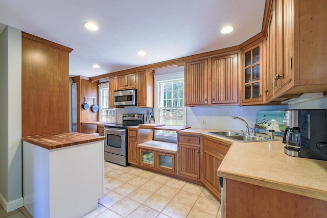 kitchen with a sink, glass insert cabinets, appliances with stainless steel finishes, brown cabinetry, and light tile patterned floors