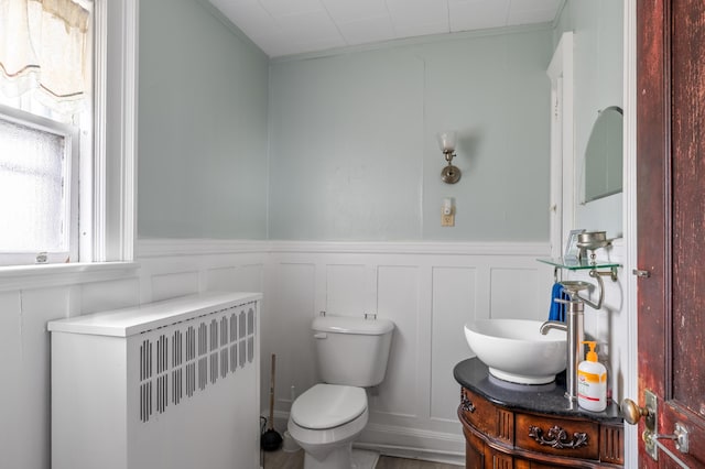 half bathroom featuring radiator, a decorative wall, vanity, a wainscoted wall, and toilet