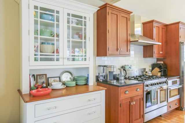 kitchen with brown cabinets, wall chimney exhaust hood, range with two ovens, and decorative backsplash