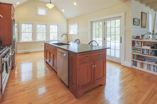 kitchen featuring light wood-style floors, appliances with stainless steel finishes, a kitchen island with sink, a sink, and dark countertops