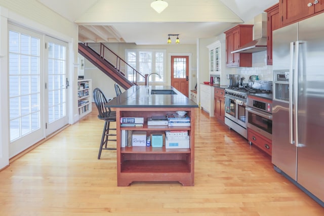 kitchen featuring wall chimney range hood, open shelves, dark countertops, appliances with stainless steel finishes, and a sink