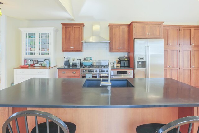 kitchen featuring a breakfast bar, dark countertops, brown cabinets, wall chimney exhaust hood, and stainless steel appliances