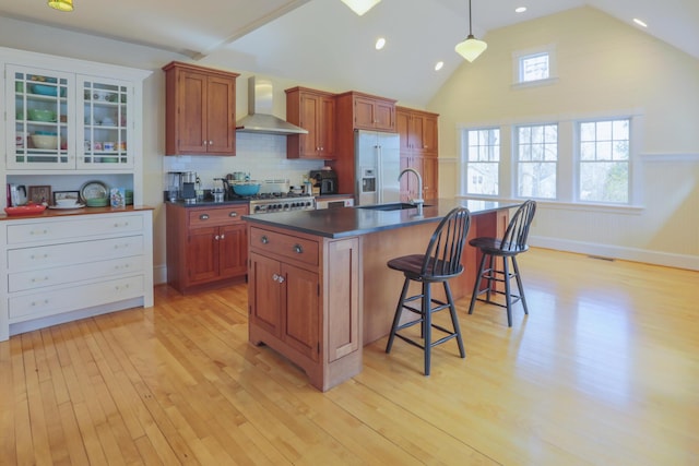 kitchen with light wood-type flooring, high end fridge, wall chimney range hood, dark countertops, and a sink