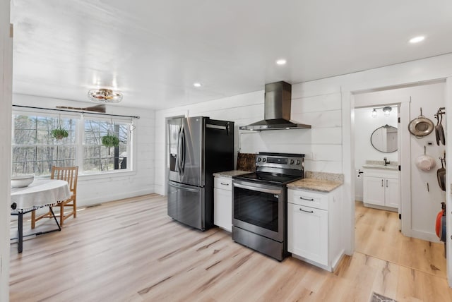 kitchen featuring wall chimney range hood, appliances with stainless steel finishes, light stone countertops, white cabinets, and light wood finished floors