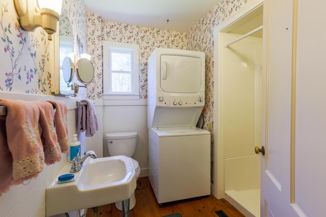bathroom featuring a sink, toilet, wallpapered walls, wood finished floors, and stacked washer and clothes dryer