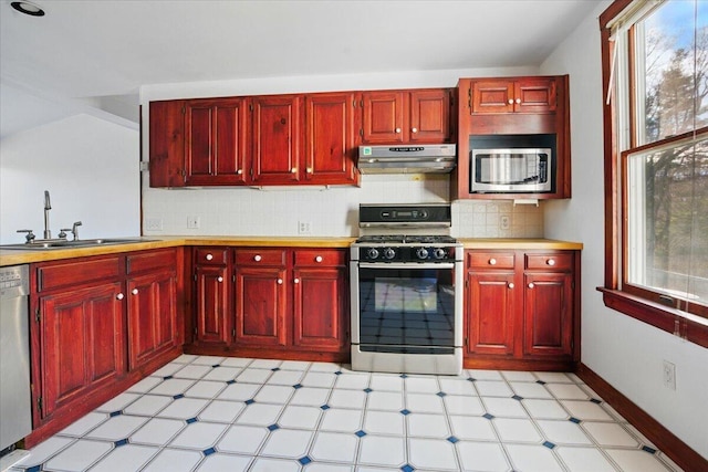kitchen with light floors, reddish brown cabinets, under cabinet range hood, a sink, and appliances with stainless steel finishes