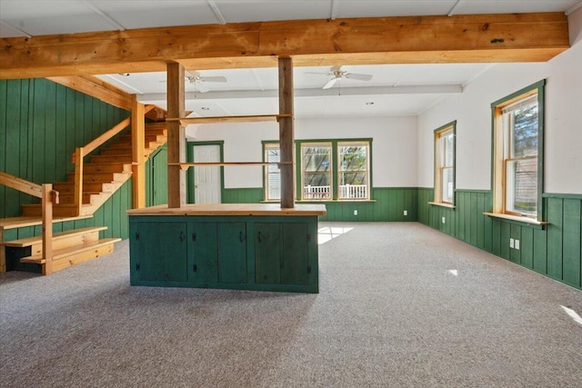 unfurnished living room with beam ceiling, carpet floors, a wainscoted wall, stairs, and a ceiling fan