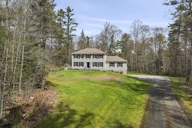colonial-style house with driveway, a front lawn, roof with shingles, a chimney, and a wooded view