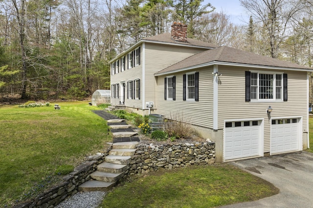 view of home's exterior with a chimney, asphalt driveway, a shingled roof, a lawn, and a greenhouse
