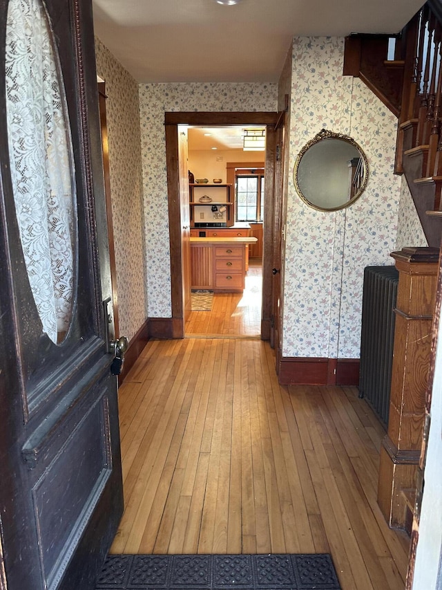 corridor featuring radiator, wallpapered walls, light wood finished floors, baseboards, and stairs