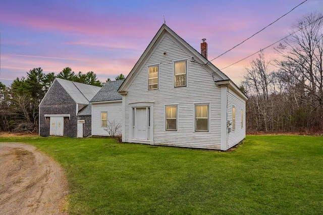 back of house at dusk featuring a chimney, driveway, a yard, and roof with shingles