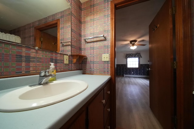 bathroom with wallpapered walls, wood finished floors, a ceiling fan, vanity, and a baseboard heating unit