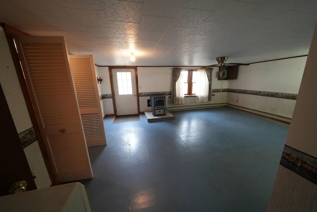 unfurnished living room featuring a baseboard heating unit, a ceiling fan, a wood stove, and a wainscoted wall