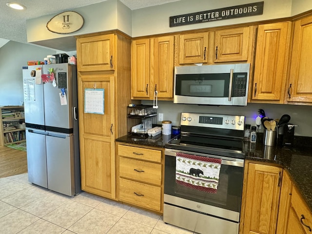 kitchen featuring brown cabinetry, dark stone countertops, stainless steel appliances, and light tile patterned floors