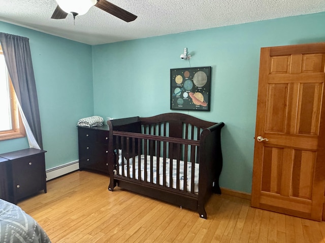 bedroom with baseboard heating, a nursery area, a ceiling fan, a textured ceiling, and hardwood / wood-style flooring