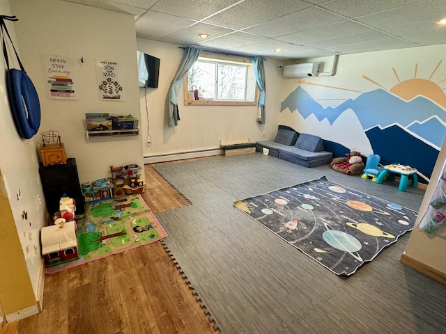 recreation room featuring a baseboard heating unit, a wall mounted AC, wood finished floors, and a paneled ceiling