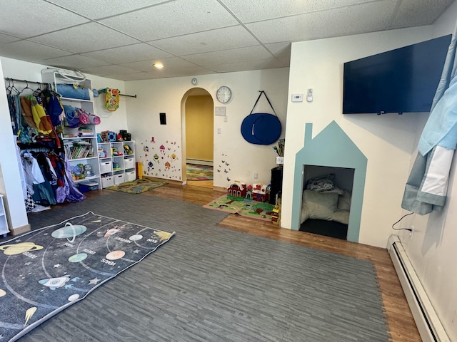 game room with a baseboard heating unit, arched walkways, a drop ceiling, baseboards, and wood finished floors