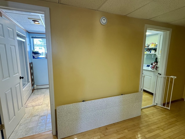corridor with a baseboard heating unit, light wood-type flooring, and a drop ceiling