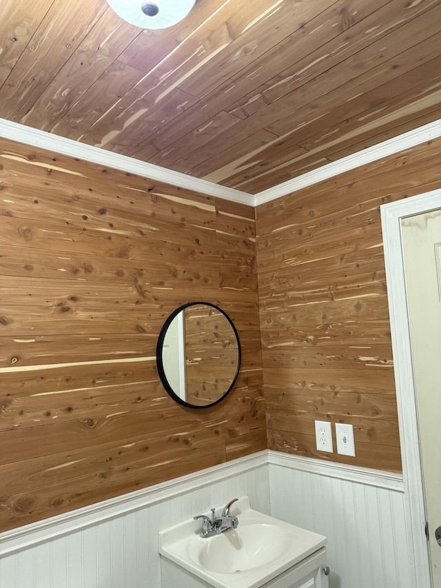 bathroom with wood walls, wooden ceiling, a sink, crown molding, and wainscoting