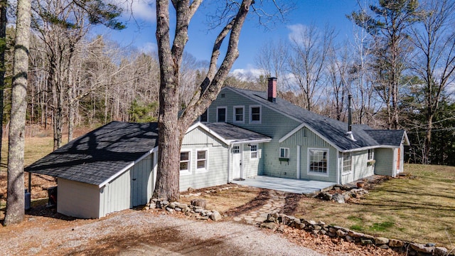 view of front facade with a chimney, a shingled roof, a patio, and an outbuilding