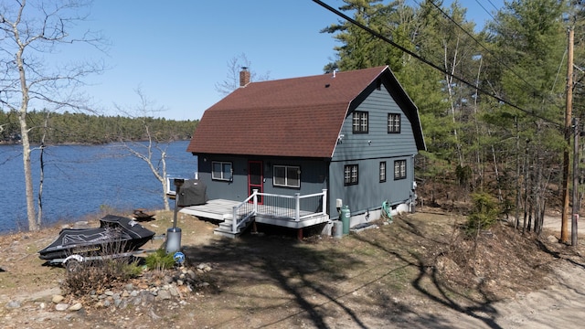 view of front facade with covered porch, a chimney, a wooded view, a gambrel roof, and a water view