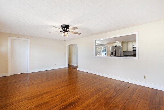 unfurnished living room with arched walkways, wood-type flooring, a ceiling fan, and baseboards