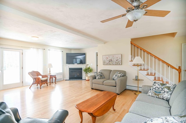 living area with a ceiling fan, baseboard heating, stairway, light wood finished floors, and a glass covered fireplace