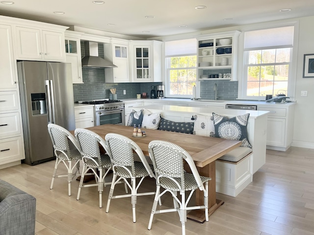 kitchen with wall chimney range hood, white cabinets, appliances with stainless steel finishes, light wood-style floors, and tasteful backsplash