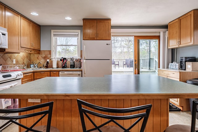 kitchen featuring a kitchen breakfast bar, backsplash, brown cabinets, a sink, and white appliances