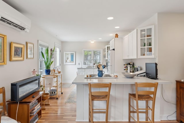 kitchen featuring glass insert cabinets, an AC wall unit, a peninsula, light wood-style floors, and white cabinetry