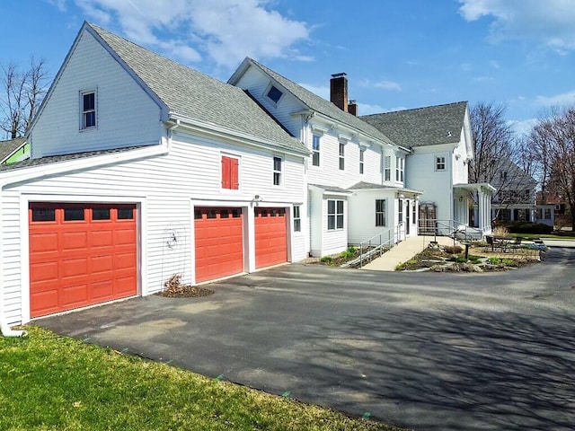 view of side of home featuring driveway, roof with shingles, a chimney, and an attached garage