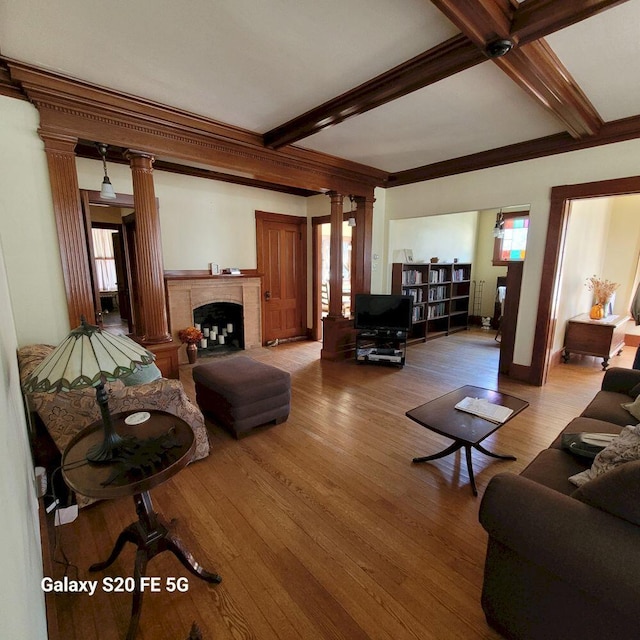 living room featuring crown molding, beam ceiling, a fireplace, ornate columns, and wood finished floors