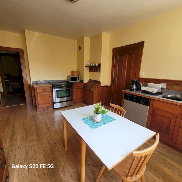 kitchen with hardwood / wood-style floors, brown cabinetry, appliances with stainless steel finishes, a wainscoted wall, and a sink