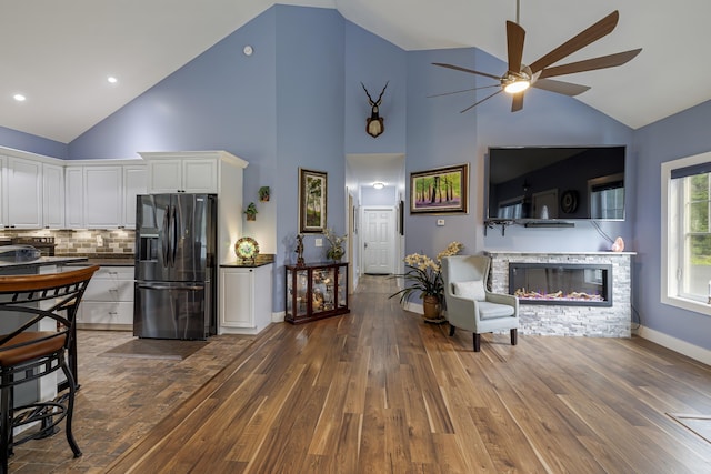 living room with baseboards, wood-type flooring, a ceiling fan, high vaulted ceiling, and a stone fireplace