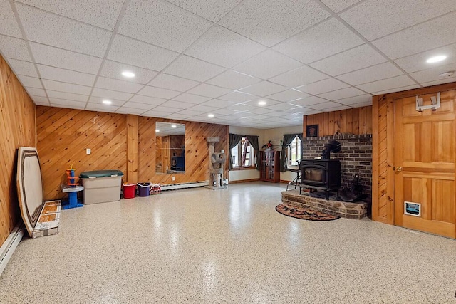 below grade area featuring a wood stove, wooden walls, a drop ceiling, and baseboard heating