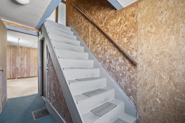 stairs with visible vents, carpet flooring, a drop ceiling, and wood walls