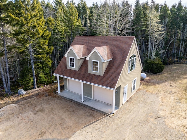 view of front facade featuring roof with shingles, an attached garage, dirt driveway, and a forest view