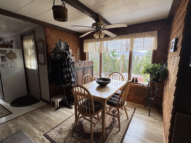 dining room with wood walls, beamed ceiling, light wood-type flooring, and ceiling fan