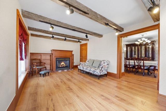 living area with light wood-type flooring, beam ceiling, track lighting, a notable chandelier, and a glass covered fireplace
