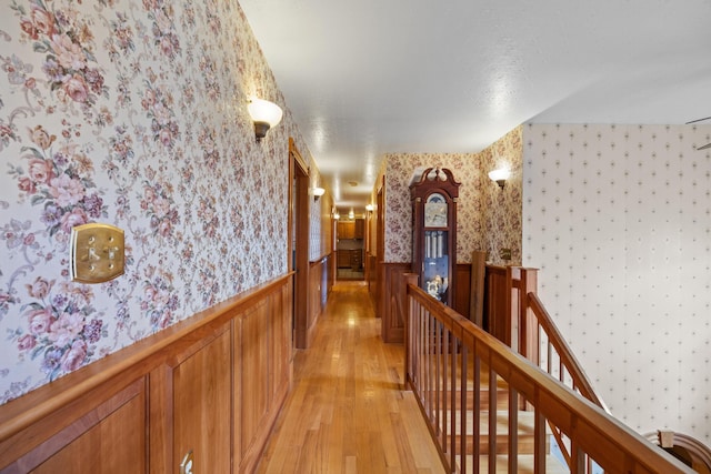 hall with a wainscoted wall, light wood finished floors, wallpapered walls, and an upstairs landing