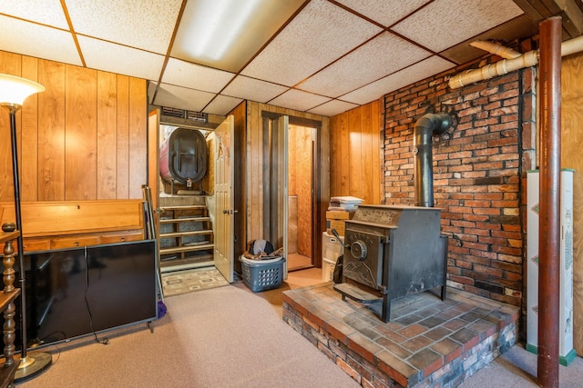 living room with a wood stove, wooden walls, carpet floors, a paneled ceiling, and stairway
