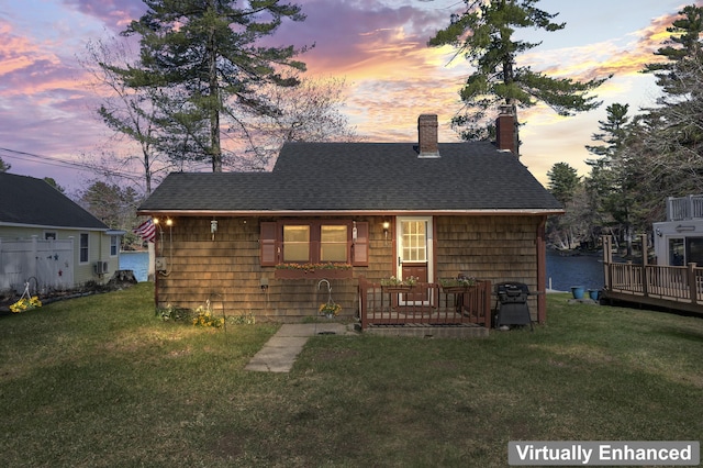 back of house at dusk with a lawn, a shingled roof, a chimney, and a deck