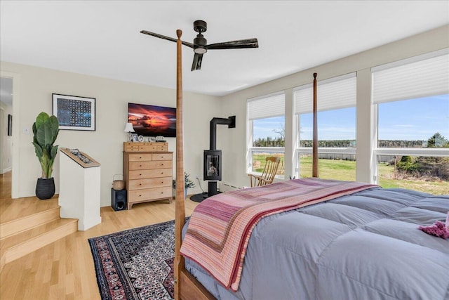 bedroom with a wood stove, ceiling fan, light wood finished floors, and a baseboard radiator