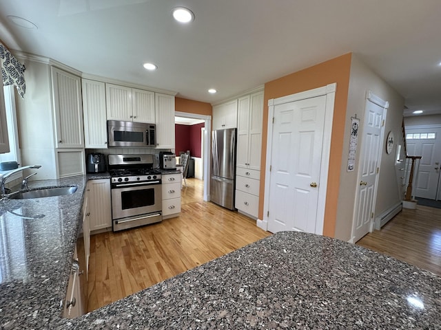 kitchen featuring appliances with stainless steel finishes, baseboard heating, a sink, dark stone counters, and light wood-type flooring