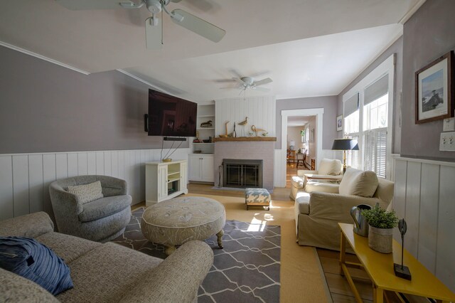 living area with crown molding, ceiling fan, wainscoting, and a brick fireplace