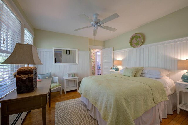 bedroom featuring wood finished floors, a ceiling fan, a baseboard heating unit, and a wainscoted wall
