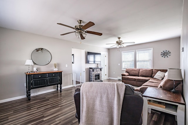 living room featuring dark wood finished floors, baseboards, a ceiling fan, and a stone fireplace